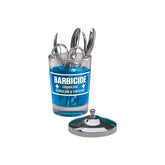 Barbicide disinfecting jar specifically designed for manicure tables or smaller salon tools. The jar is filled with a bright blue liquid and holds several pairs of metal salon shears, nippers, or tweezers. The iconic blue and white Barbicide label is clearly visible on the front, featuring the words "Germicide, Fungicide & Virucide." The stainless steel lid and internal self-draining retrieval basket assembly are resting flat on the surface next to the base of the glass jar. The entire product image is cent