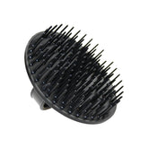 A circular, all-black scalp massage and shampoo brush. The brush features a dense array of short, flexible plastic bristles on its face and a molded finger loop handle on the back for a secure grip. It is shown at a three-quarter angle against a plain white background.