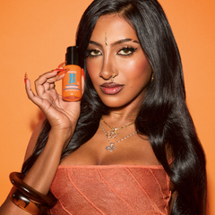 A woman with long black hair holds up an orange bottle of Nutra Body Butter against a solid orange background.