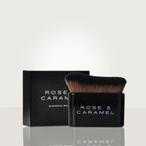 A Rose & Caramel blending brush with black bristles and a black handle, shown in front of its matching black packaging box on a light gray reflective surface.