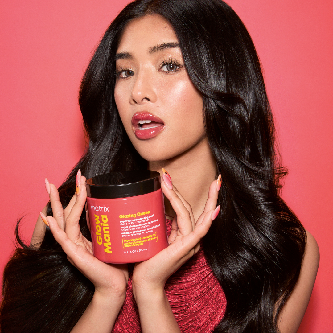 A woman with long, wavy black hair holds a red tub of Matrix Glow Magic hair mask against a solid pink background.