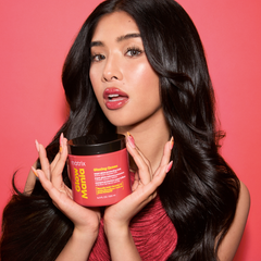 A woman with long, wavy black hair holds a red tub of Matrix Glow Magic hair mask against a solid pink background.