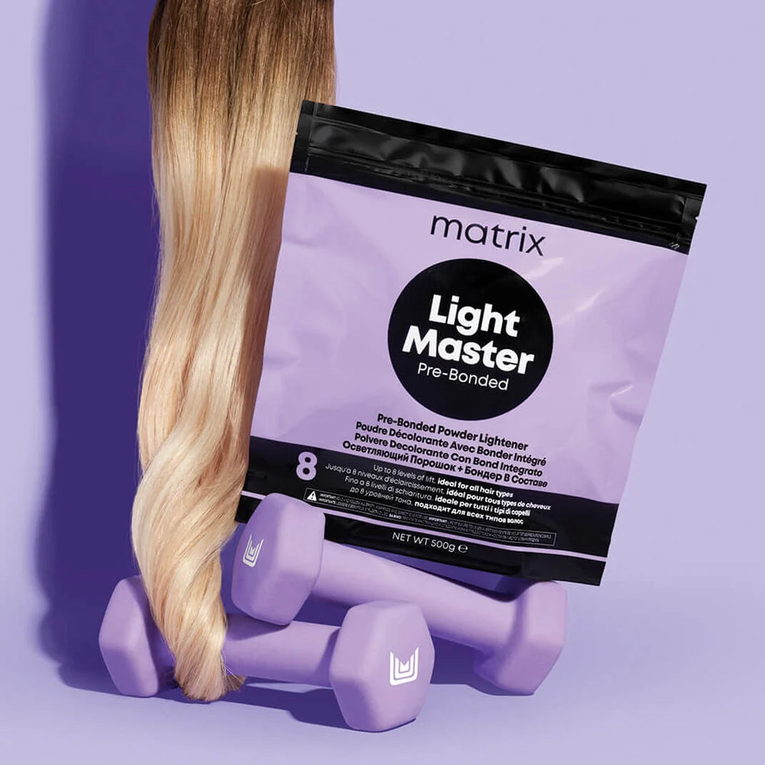 A bag of Matrix Light Master Pre-Bonded powder lightener in shade 8, styled with a long blonde hair extension and two lavender dumbbells on a matching lavender background.