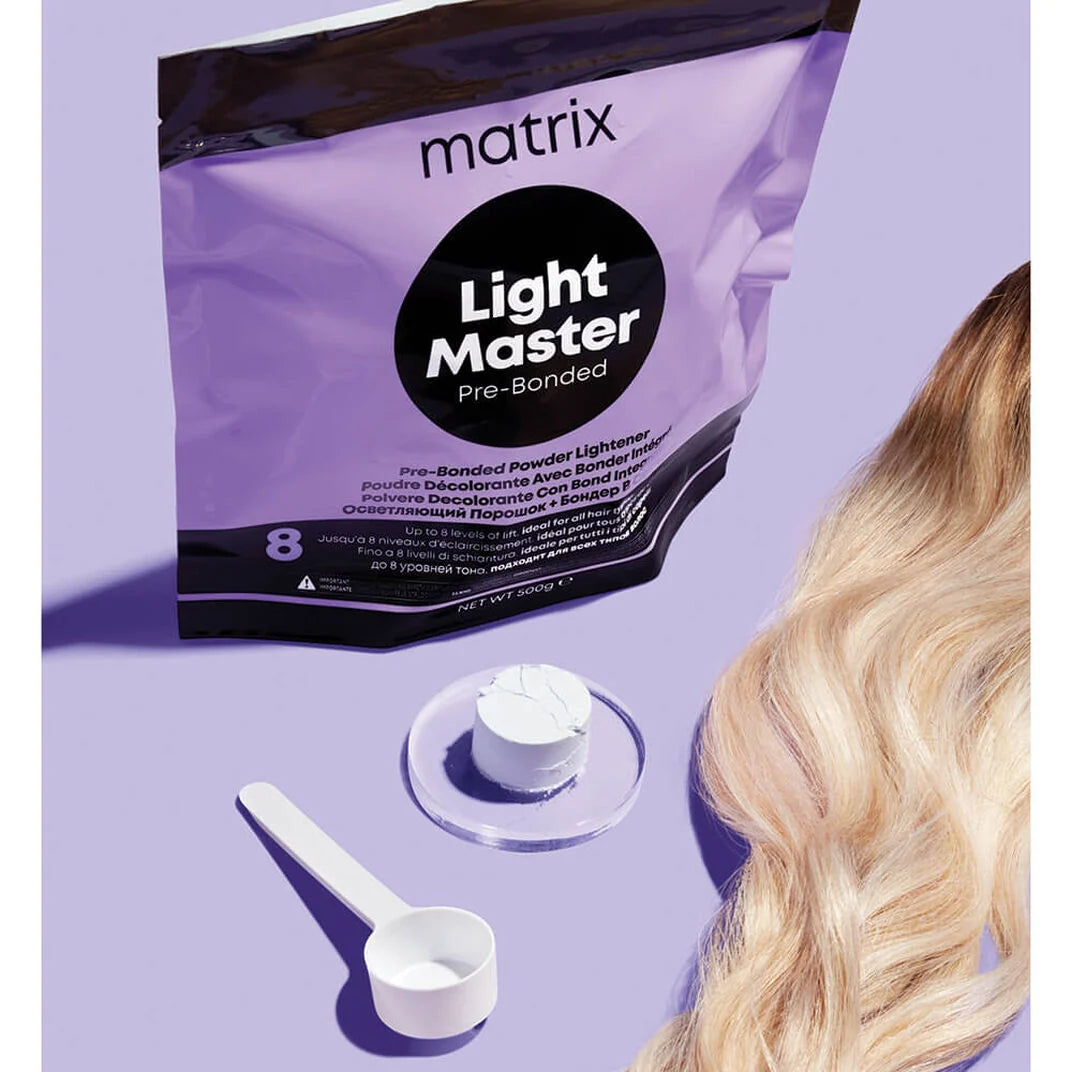 A purple Matrix Light Master Pre-Bonded powder package with a white scoop and powder dish, shown beside long blonde hair on a lavender background.