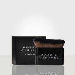 A Rose & Caramel blending brush with black bristles and a black handle, shown in front of its matching black packaging box on a light gray reflective surface.